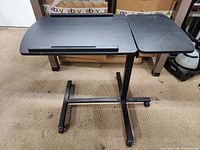 Side view of the black rolling adjustable height table showing the two tabletop surfaces and metal frame with wheels.