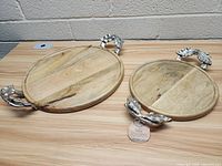 Two round wooden serving trays with silver leaf-shaped metal handles, one larger and one smaller, shown on a wooden table against a concrete block wall.