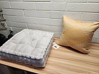 Photo showing super thick beige tufted chair cushion with yellow striped throw pillow on a wooden surface against a brick wall