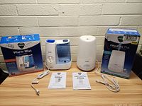 Two Vicks humidifiers placed on a table; Warm Mist on the left with box, FilterFree Plus on the right with box; instruction manuals and cords in front; displayed to show all items included in the lot.