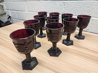 Set of nine hexagonal stemmed red glass goblets arranged on a wooden surface with white brick wall background.