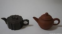 Photo showing both teapots side by side on white background, black carved and brown clay.