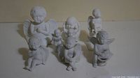 Overview photo showing all seven white cherub angel figurines arranged on a flat surface, varying in poses and sizes.