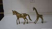 Photo showing both bronze horse and giraffe figurines side by side on a white surface with tiled wall background.