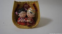 Front angled view of a figurine set showing the bride and groom in traditional Chinese wedding attire seated inside a textured basket frame decorated with Chinese red symbols for happiness.