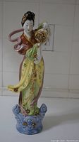 Full view of the Japanese porcelain figurine showing the woman standing on a blue wave-like base, wearing a red kimono top and yellow-green skirt with floral patterns, holding a gold ornate lantern.