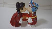 Full view of both bride and groom porcelain wedding figurines kissing and holding a red pillow with gold embroidery.