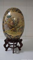 Front view of the porcelain egg, showing floral pattern in gold and colorful tones on beige background and dark blue border at the top with small gold designs, placed on wooden stand.