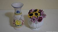Pair of small decorative floral ceramic vases showing front view, one taller with yellow flower and painted design, one shorter with multi-colored sculpted floral bouquet on top.