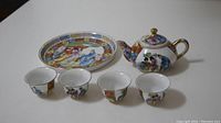 Photo showing entire tea set including teapot, four cups, and one plate, all decorated with colorful Asian-inspired figures and floral designs.