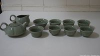 Full tea set laid out, showing 1 teapot with lid, 1 creamer or small pitcher, 1 large cup, and 7 small cups in celadon green ceramic.
