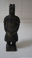 Front view of terracotta warrior figurine showing its full body with detailed armor and hands clasped at front.