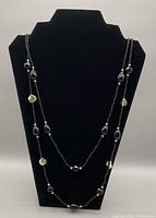 Image of long necklace featuring black beads and gold-tone dangle tassels displayed on a black stand.