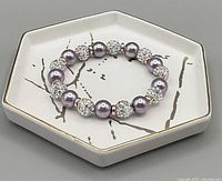 Lilac colored iridescent bead and rhinestone bead bracelet displayed on hexagonal dish with gold rim and bird branch design.