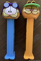 Front view of two Garfield Pez dispensers with distinct headgear and stem colors, placed side by side on a wooden surface.
