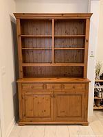 Front view of wooden buffet and hutch showing natural wood finish, two cabinet doors, three drawers, and hutch with shelves.
