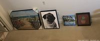 Four prints on the floor against a wall in the basement, showing overall arrangement and size comparison including a beach scene, pug portrait, motivational quote, and succulent print.