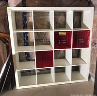 Full view of white 16-cube shelving unit with three red bins inserted in cubbies, against unfinished basement wall.