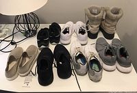 Overview of 8 pairs of shoes including beige boots, white sneakers, black shoes, grey sneakers, and slip-on shoes laid out on a white table.