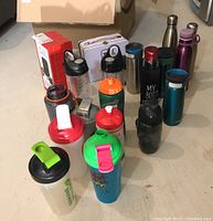 Photo showing assortment of travel mugs, water bottles, metal lunch box, and hot cocoa kit boxed items on the ground.