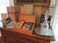 Photo showing wooden box with bartenders guide books, Vieux Marc glass decanter and matching cup, cut glass decanter, metal bottle carrier, and other barware on wood surface.