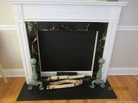 Full view of the portable faux fireplace showing the white painted surround with decorative motifs, black backing panel, faux logs, and green metal andirons placed on a black base.