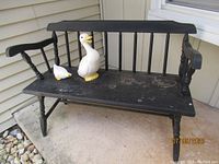 Full view of the black painted wooden garden bench showing one loose armrest and two ceramic duck figurines placed on the bench seat