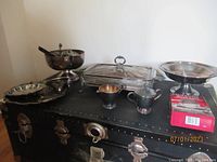 Lot of silver plated serving pieces including Fire-King glass casserole dish with silver plated lid and stand alongside other silver plated serveware on black surface.