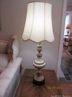 Full view of one lamp lit on wooden side table next to cream sofa, showing off scalloped fabric lampshade and brass and ceramic lamp base on black base.