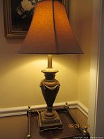 Table lamp lit, showing the warm glow of the fabric shade and the urn-shaped base with gold and brown tones and decorative motifs.