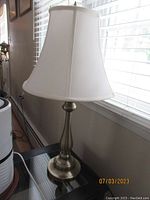 Single brushed metal table lamp with white shade placed on glass table beside window.