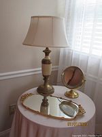 Photo of the vintage ormolu vanity/tray mirror, Jerdon magnifying mirror, and table lamp on a small round table by a window.