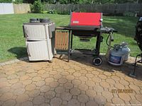 Wide-angle photo showing Weber red-lid gas grill, two gas canisters, and adjacent beige Suncast cabinet outdoors on patio.
