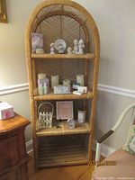 Front view of vintage wicker bookshelf with items displayed on three shelves including ceramic figurines, specialty soaps, Lenox trinket box, candles, picture frame, and decorative birdcage.