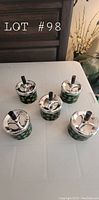 Five identical ashtrays with a shiny chrome push-down lid and green leaf-patterned bodies on a table.