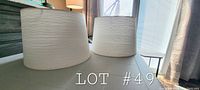 Two cream color fabric lampshades sitting on a table, showing their cylindrical shape and textured horizontal fabric pattern.