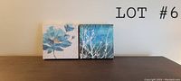 Two square canvas art pieces side by side on a floor against a wall, showing a blue flower and white branches on blue background; Lot #6