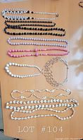 Top-down photo showing seven necklaces in various bead colors and styles including grey, black, white, pink, and silver-tone chain link necklace arranged on a wooden surface.