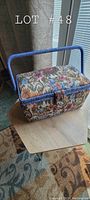 Front and side view of rectangular floral fabric sewing kit case on wooden table, showing colorful flowers and blue woven handle.