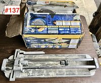 Two Rubi TX-1000 tile cutters including one with original box shown from top view on a dark surface, visible signs of wear and dust.