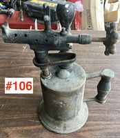 Front angle view of the antique brass blow torch showing the cylindrical fuel tank, pump handle, valve wheel, and nozzle.