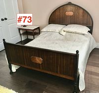 Full view of antique wooden double size bed with white quilted bedspread and two pillows in bedroom setting near bedside table.