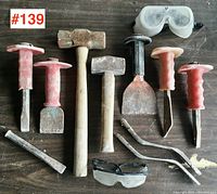 Top-down view of the full lot of stonework chisels, hammers, pry bars, and safety glasses arranged on a dark surface.