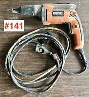 Close-up image of Ridgid drywall screwdriver with attached power cord placed on a wooden surface. The tool and cord show signs of use and dirt.