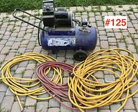 Blue Campbell Hausfeld portable air compressor on wheels with handle, sitting on stone patio, with three coiled air hoses in yellow and red in front.