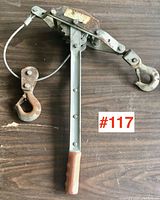 Top-down view of ratchet cable tool with visible handle, cable, and hooks, showing rust on hooks.