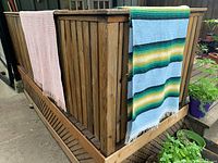 Two woven textiles draped over a wooden railing side by side, showing carpet with colorful stripes and pink blanket.