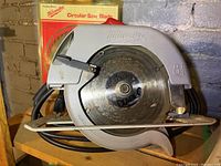 Front view of Milwaukee circular saw showing blade, guard, and power cord with extra blade in original packaging in background.