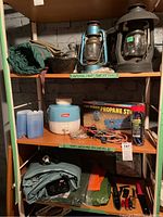 Wide view of three shelves showing lanterns including vintage Dietz oil lanterns, Yanes propane stove box, Coleman water cooler and camping gear like tarps and bungees.