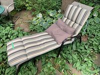 Patio lounger with striped cushions and two throw pillows positioned on garden ground with greenery around